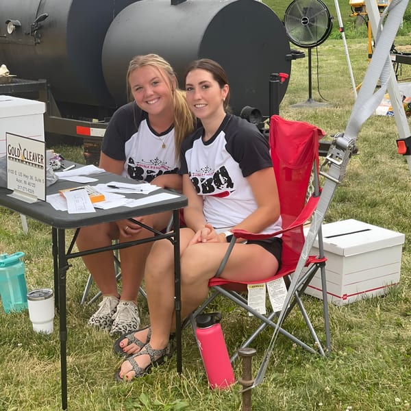 Two smiling women in Double D's Southern BBQ shirts sit at a table outdoors with a large smoker behind them.