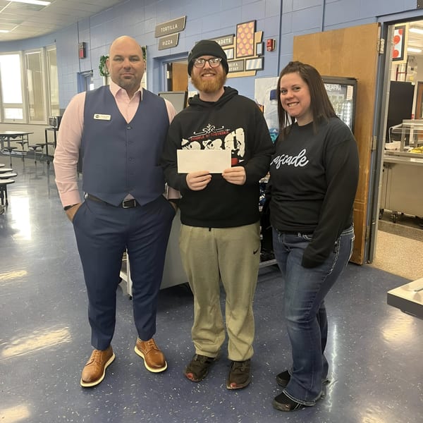 Three people, two men and one woman, stand smiling in a school cafeteria, one holding an envelope.