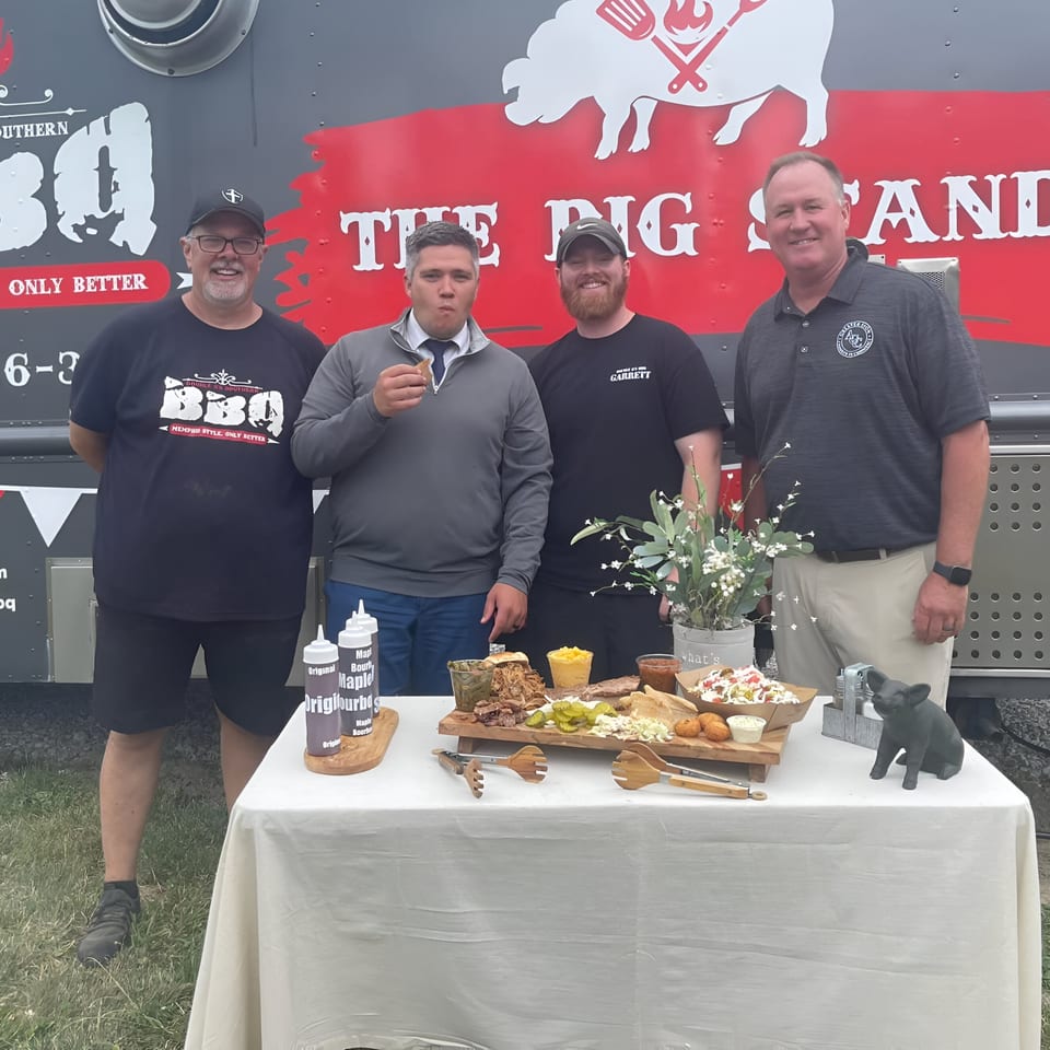 Four men stand by a Double D's Southern BBQ food truck with a table of BBQ dishes.