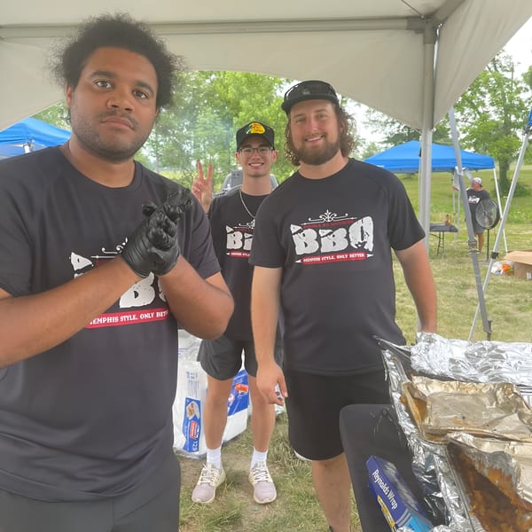 Three smiling men in Double D's Southern BBQ shirts stand under a tent with foil-covered food.